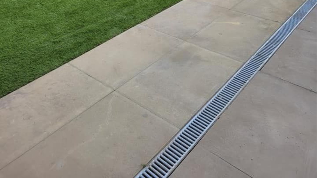 A stone patio walkway runs alongside a patch of green grass, with a long metal drainage grate embedded in the pavers.