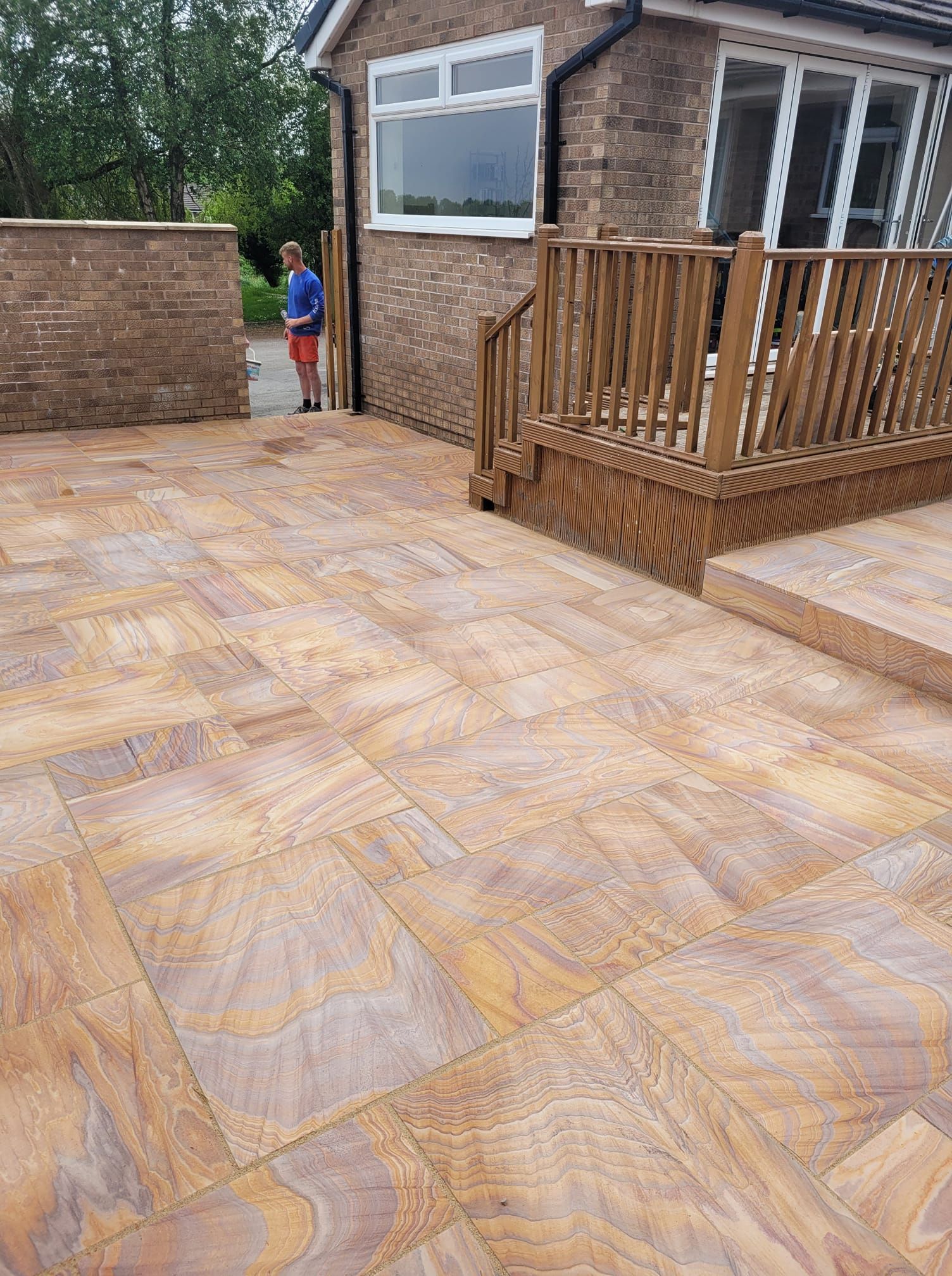 A person in a blue shirt standing near a newly laid patio of multi-colored sandstone pavers beside a wooden deck.