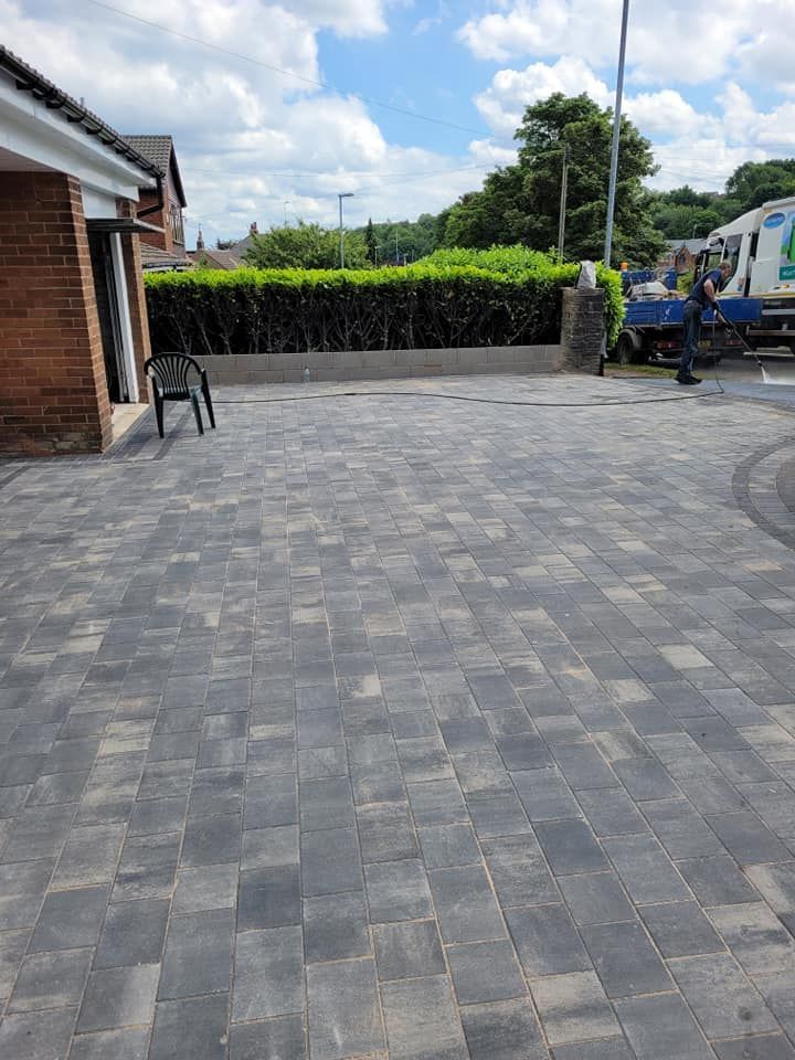 Newly laid grey block paving driveway leading to a brick house, next to a green hedge and parked utility truck.