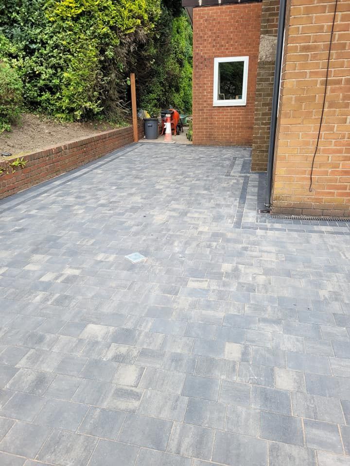 A newly laid paved driveway with gray rectangular stones, bordered by a low brick wall and the side of a brick house.