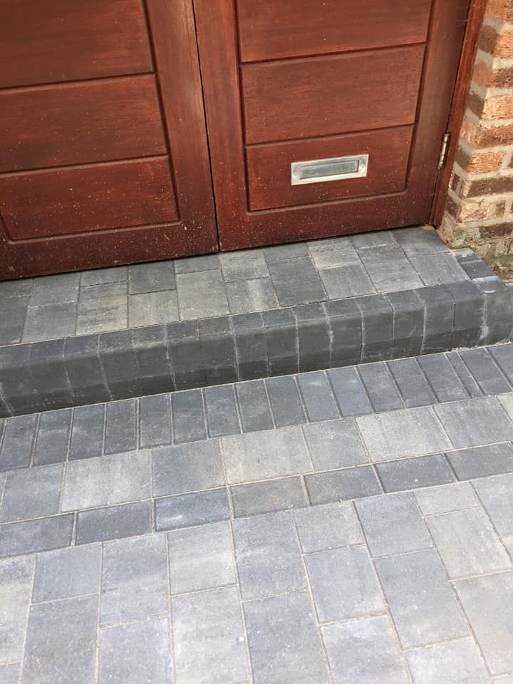A single-step entrance featuring grey paving stones with a darker border, leading up to a wooden double door.