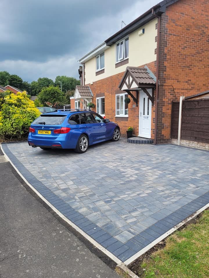 A blue station wagon is parked on a newly paved gray driveway in front of a brick two-story suburban house.