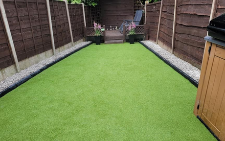 A narrow backyard garden with artificial green turf, bordered by light grey stones and enclosed by dark wooden fencing.