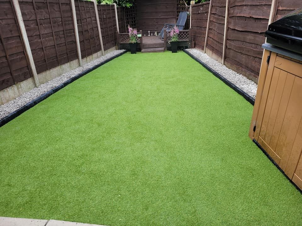 A rectangular backyard lawn with artificial green turf, bordered by decorative stones, wooden fences, and a patio area.