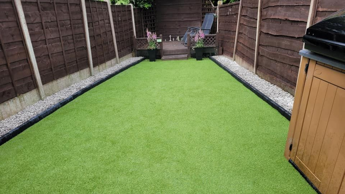 A rectangular garden with artificial green turf, bordered by gravel paths, dark wooden fences, and a wooden outdoor unit.