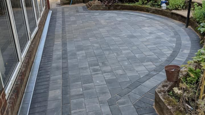 A curved grey stone patio with a dark border, adjacent to a house with glass doors and a drainage channel.