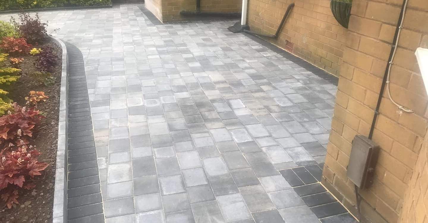 A residential driveway paved with grey rectangular stones, bordered by dark charcoal bricks and a side flower bed.