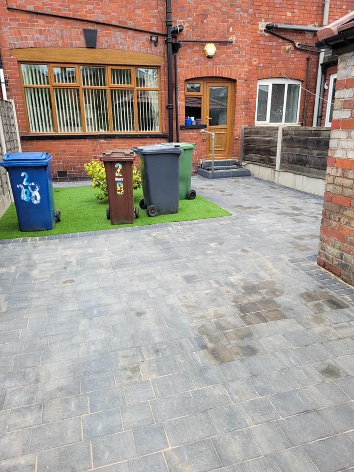 A paved courtyard with three colorful wheelie bins sitting on a small patch of artificial grass against a brick building.