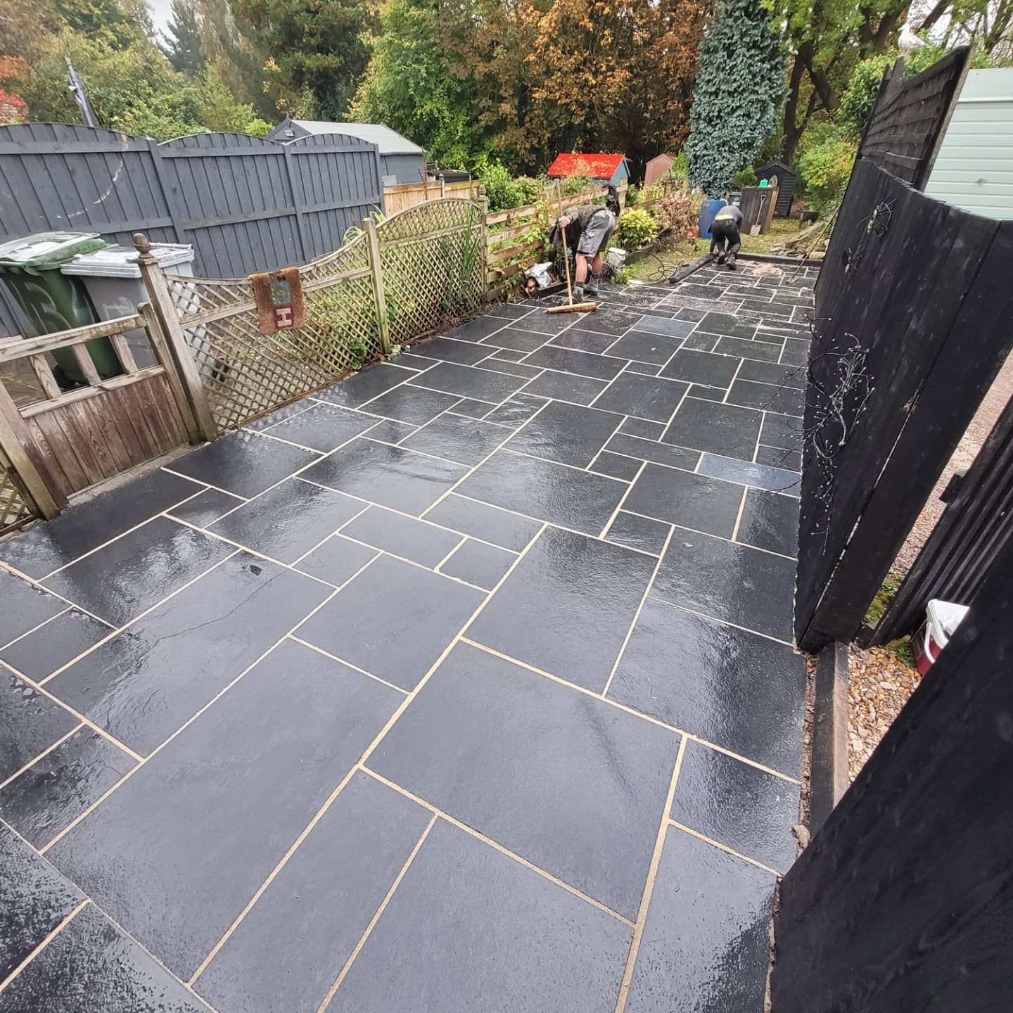 A newly laid dark grey patio with light grout lines in a backyard, with two workers cleaning the surface nearby.