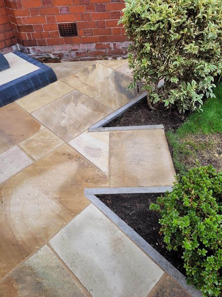 A stone patio with a zigzagging edge separating pavers from two triangular garden beds near a red brick wall.