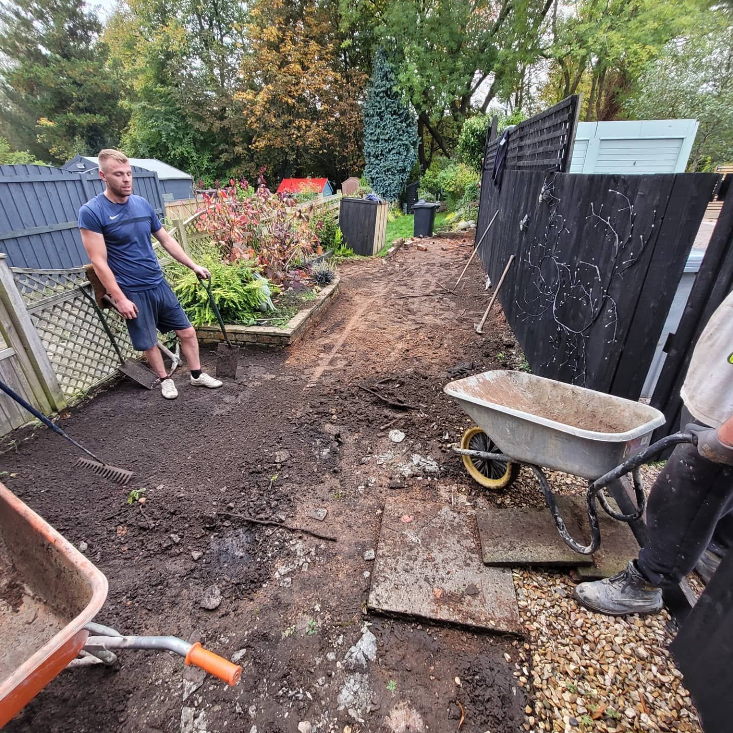 Two people clear dirt in a residential backyard with wheelbarrows and tools near a black fence and garden bed.