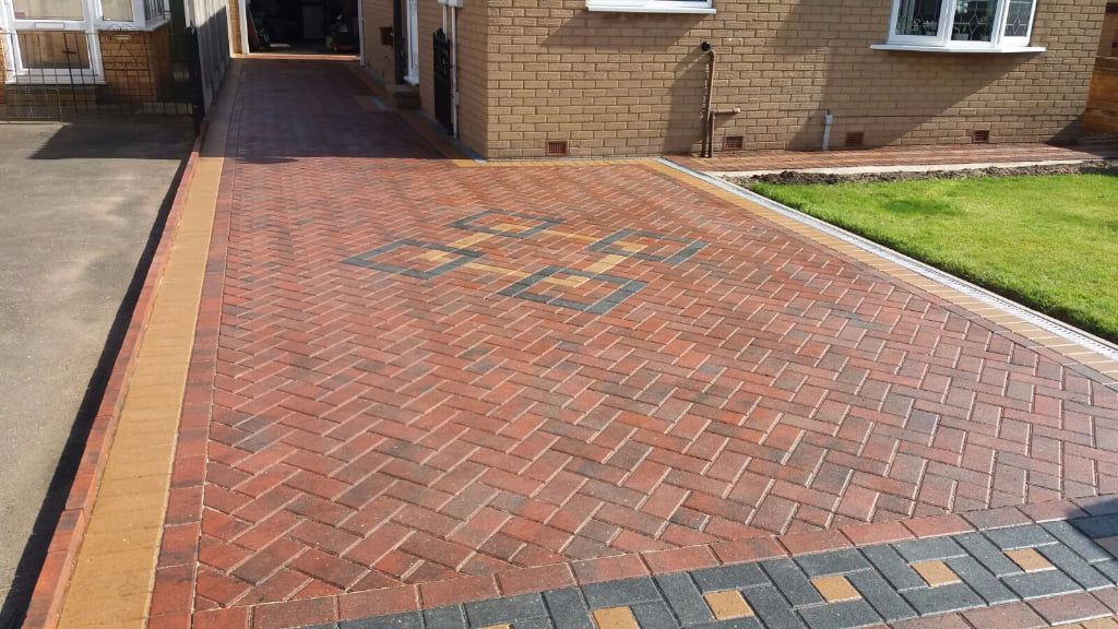 A paved driveway featuring a herringbone brick pattern with decorative border details and geometric inlays.