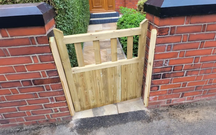 A light-colored wooden garden gate installed in a red brick wall gap, leading to a path with stone steps.