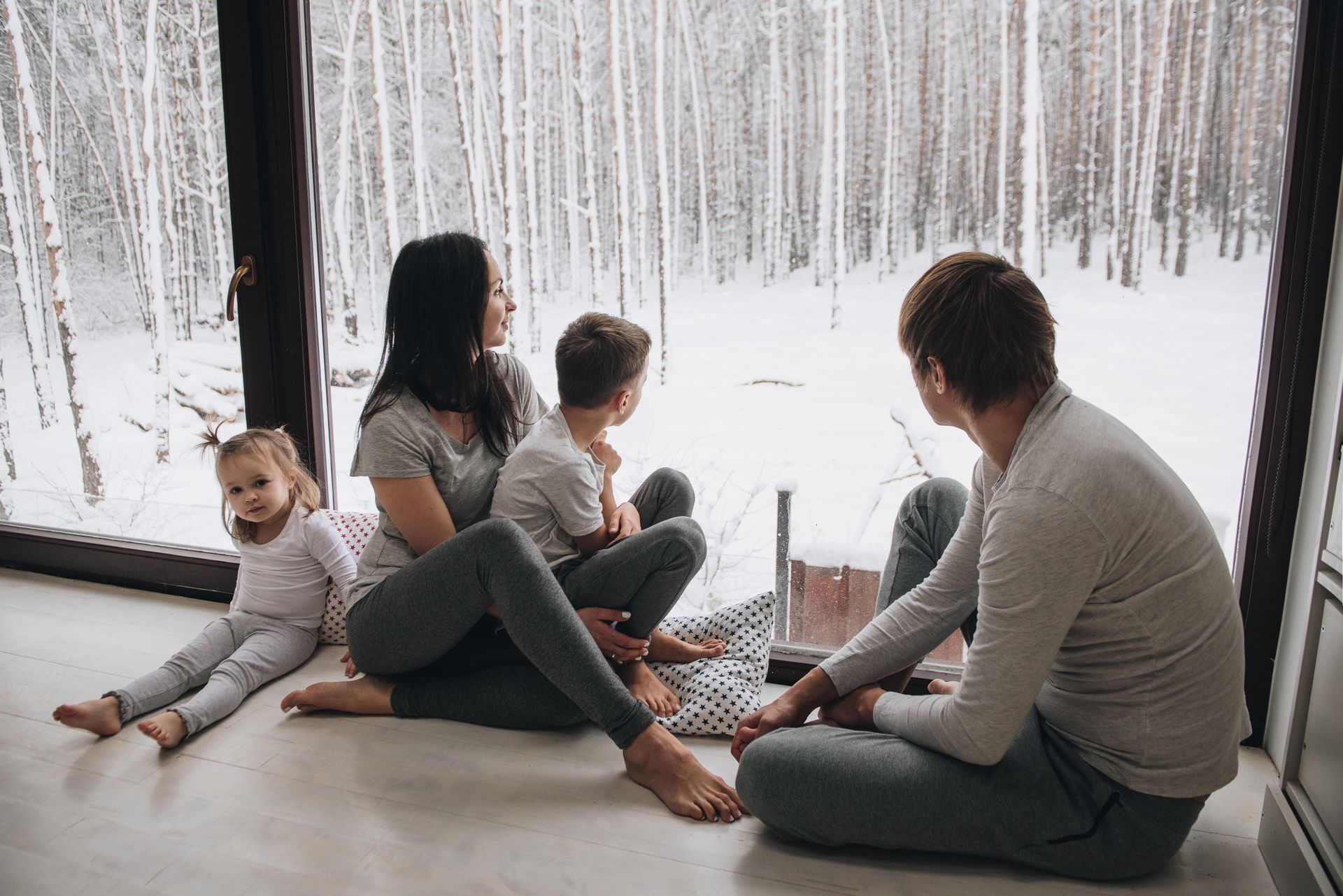 A family sitting, looking outside their expansive window, in need of covering, enjoying the snowy la