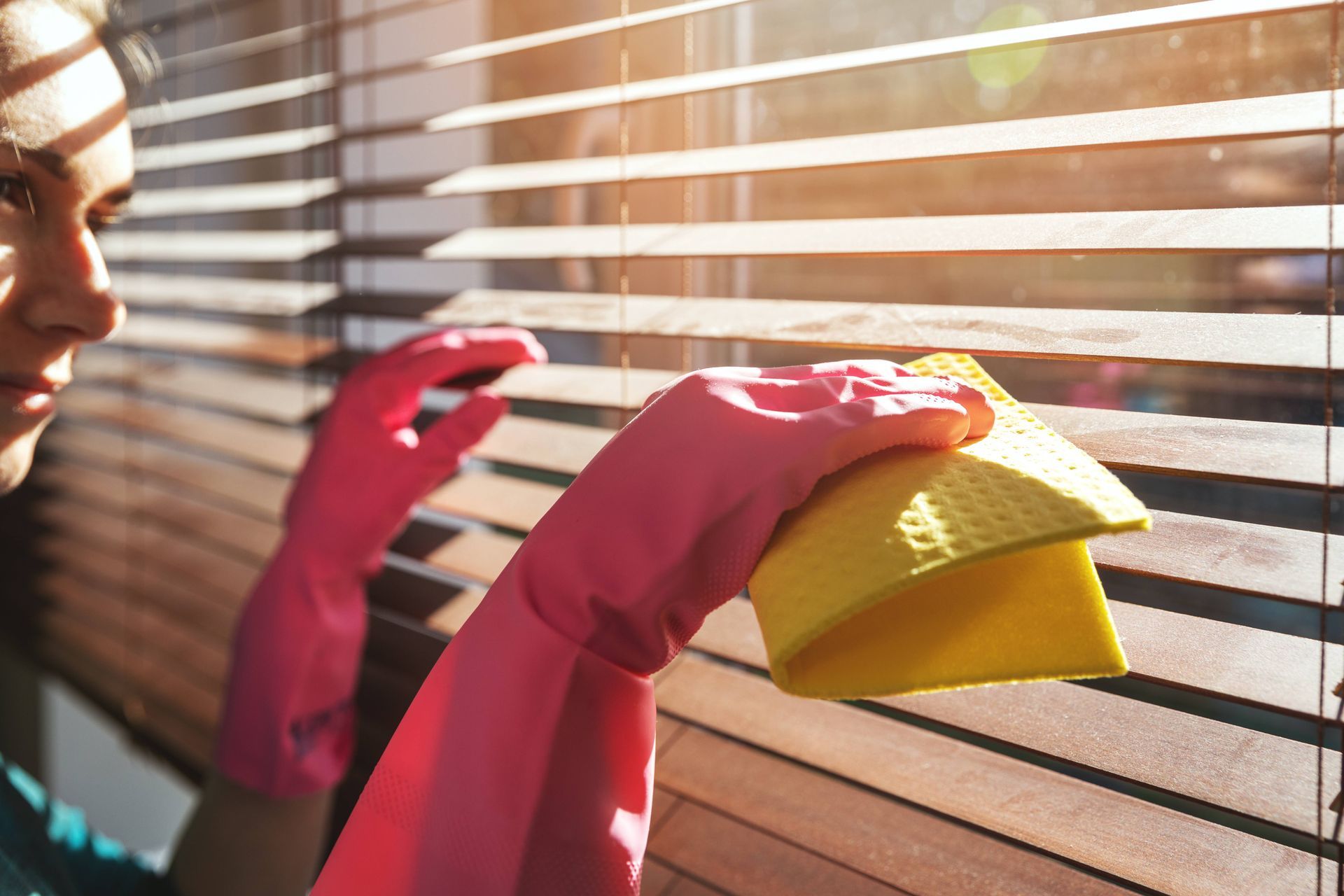 A person wearing pink cleaning gloves, using a yellow microfiber to clean their wood blinds near Anc