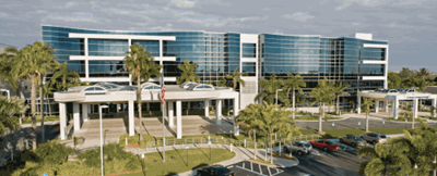 Modern office building with glass facade, palm trees, and parking area.