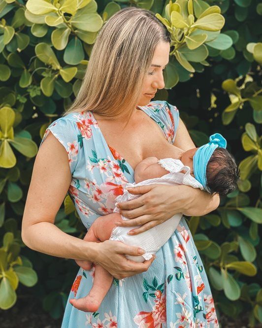 Woman in floral dress breastfeeding a baby outdoors; green foliage in background.