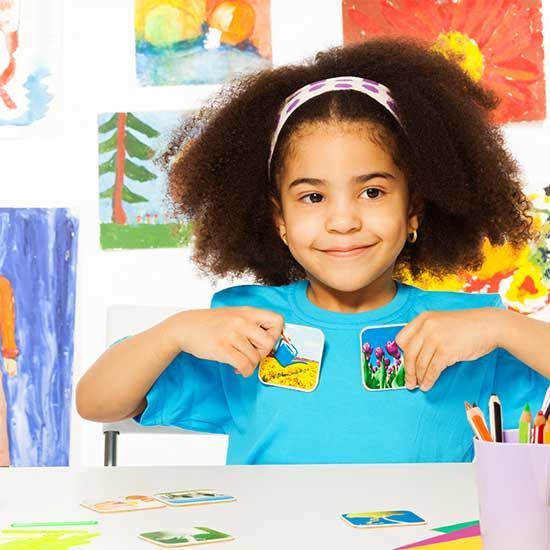Girl holding up two small paintings at a desk, smiling. Colorful artwork on wall.