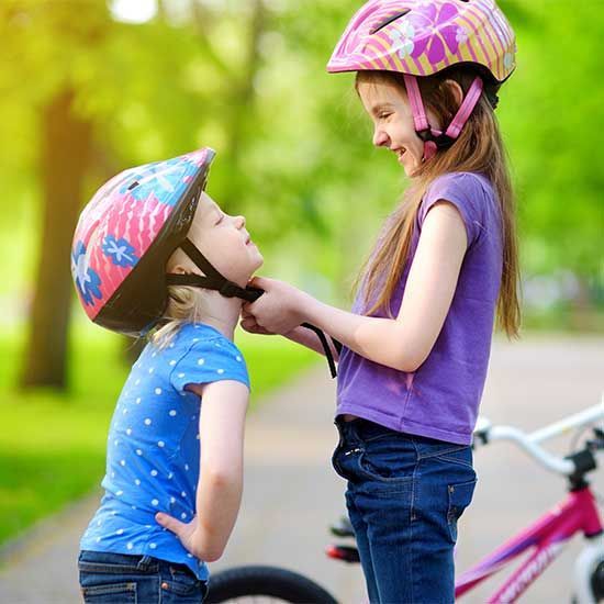 Two girls in bike helmets, one adjusting the other's helmet strap; outdoors with a bicycle.