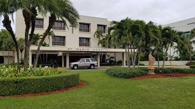 St. Mary's Hospital building with a vehicle parked in the entrance, lush green lawn, palm trees.