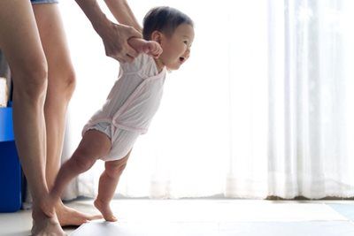 Baby learning to walk, held by an adult near a window; smiling, bright light, neutral tones.