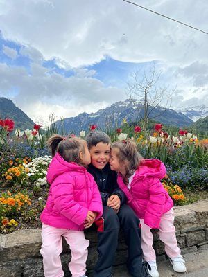 Three children in pink jackets and pants, kissing a boy seated, in front of flowers and mountains.