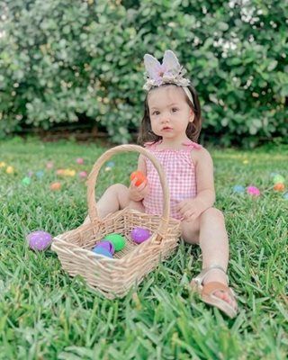 Child in gingham swimsuit and bunny ears sits in grass, holding Easter basket and egg.