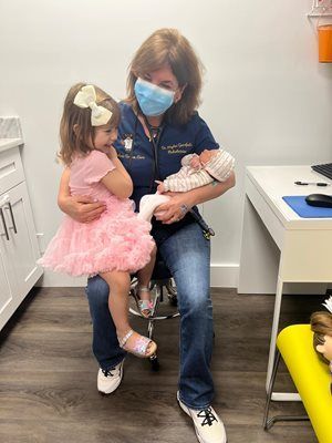 Doctor holding a baby and toddler in exam room. Both children are looking at the doctor. The doctor wears a mask.