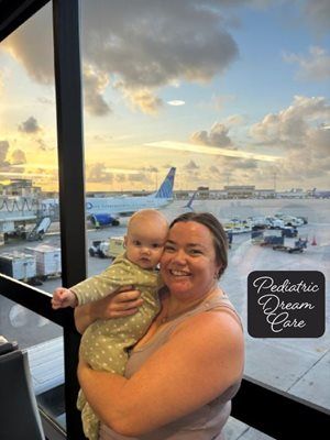 Woman holding a baby at an airport window; plane in background, sunset colors, text 