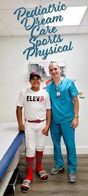 A boy in a baseball uniform smiles next to a healthcare provider in scrubs. They are in a medical setting.