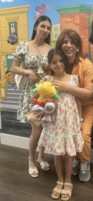 Three people smiling: a child holding a toy and two adults. They stand in front of an Sesame Street mural.