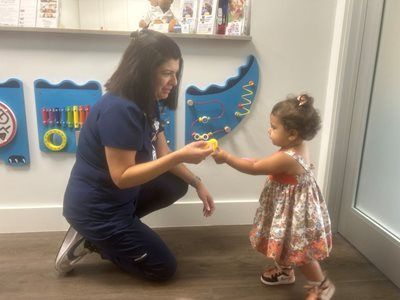 Nurse handing a yellow toy to a young child in a brightly lit room with wall-mounted toys.