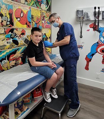 A healthcare worker gives a vaccine to a boy in a room with comic-themed wallpaper.