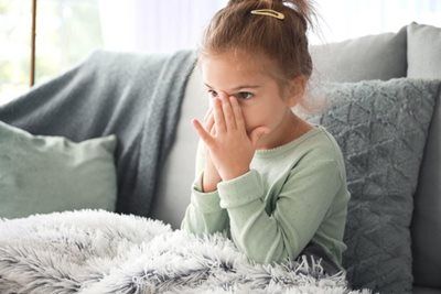 Girl with hands covering eyes, sitting on a couch with a blanket.