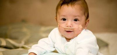 Baby with a nasal cannula smiles. Wearing a white onesie, on a bed, looking at the viewer.