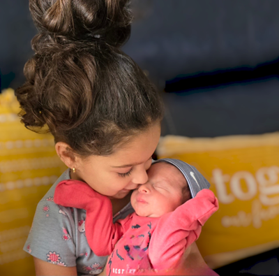 Older child holding and kissing a newborn, both in pink clothing.