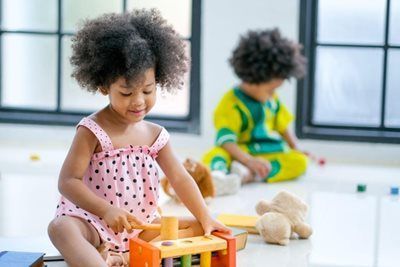 Girl in pink dress playing with wooden toy; another child in yellow shirt in the background.