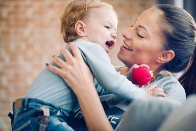 Woman smiling while holding and playing with a baby. Baby's mouth is open; red toy in its hand.