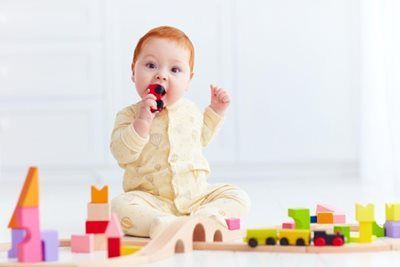 Baby with red hair sits on floor, playing with colorful wooden train and blocks, chewing on a toy.