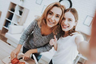 Woman cuts vegetables, smiles with girl taking selfie in a kitchen, thumbs-up.