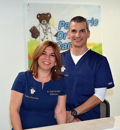 Two medical professionals smiling, standing in front of a Pediatric Dr. Care sign. They wear blue scrubs.