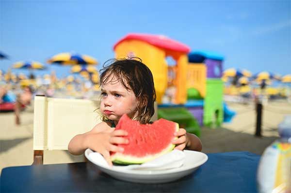Child eating watermelon at a beach, seated at a table. Brightly colored playground and umbrellas in background.