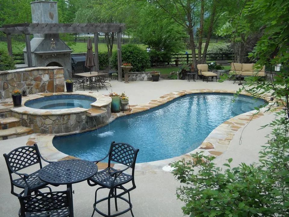 A backyard pool and hot tub surrounded by stone, with an outdoor fireplace and patio furniture.