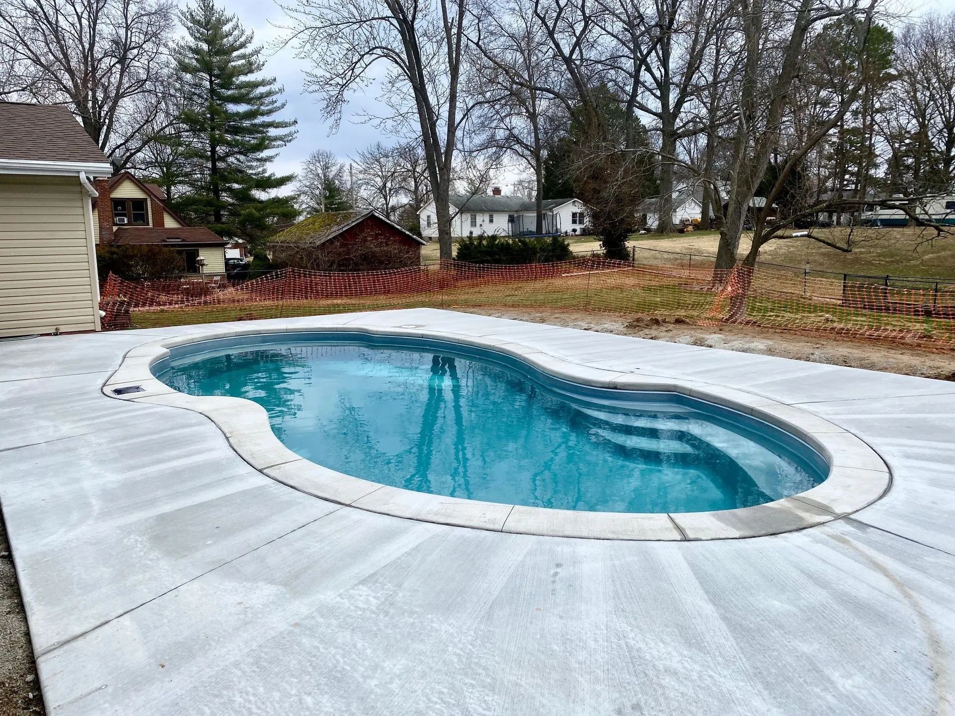 A kidney-shaped swimming pool with blue water and concrete decking. Trees and houses are
in the back