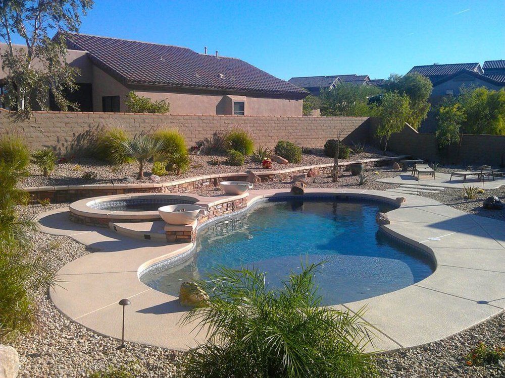 A backyard swimming pool with an integrated spa, stone landscaping, and a patio, set against a house and desert shrubs.