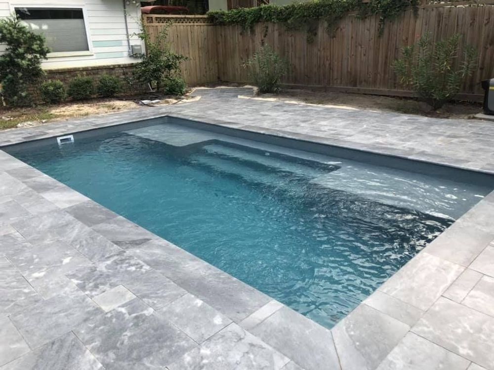 Rectangular blue swimming pool with steps, surrounded by gray stone patio in backyard.