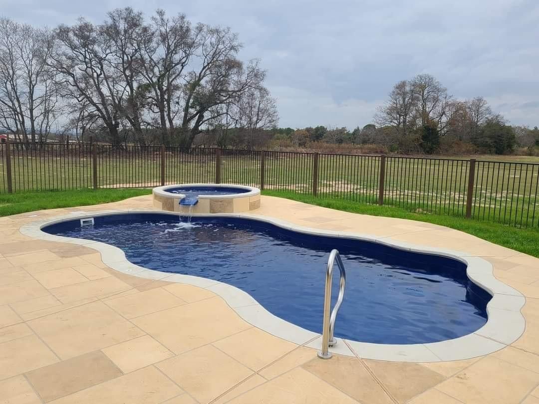 An organically shaped blue swimming pool and attached hot tub with a neutral-colored stone patio, surrounded by a metal fence.