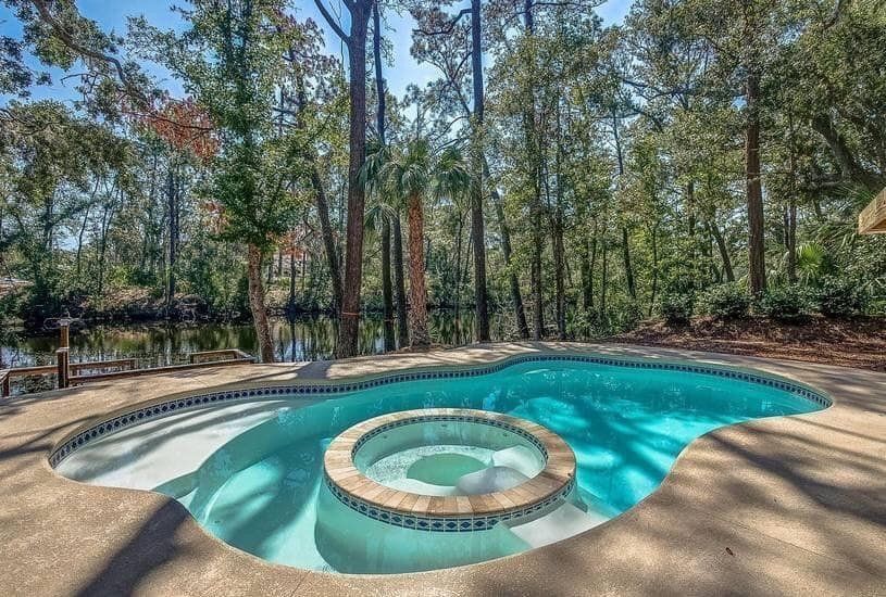 A large swimming pool with a hot tub in the middle is surrounded by trees.