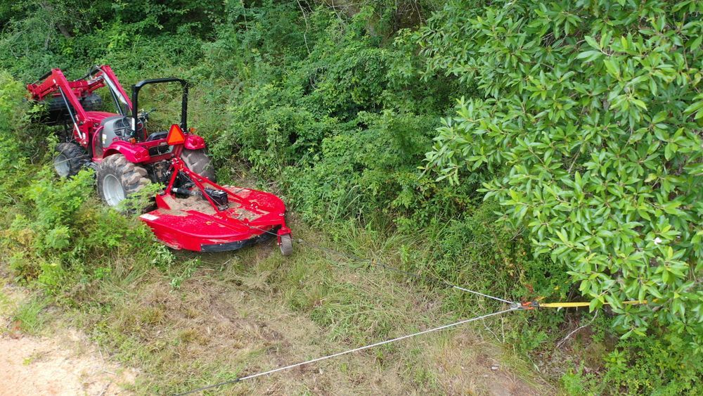 Red brush cutter in a ditch beside dense green shrubs, with a cable stretched across the slope.