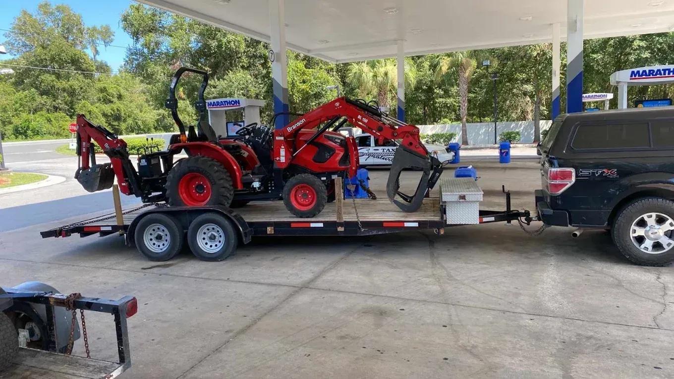 Red tractor with loader and trailer parked under a gas station canopy beside a pickup truck